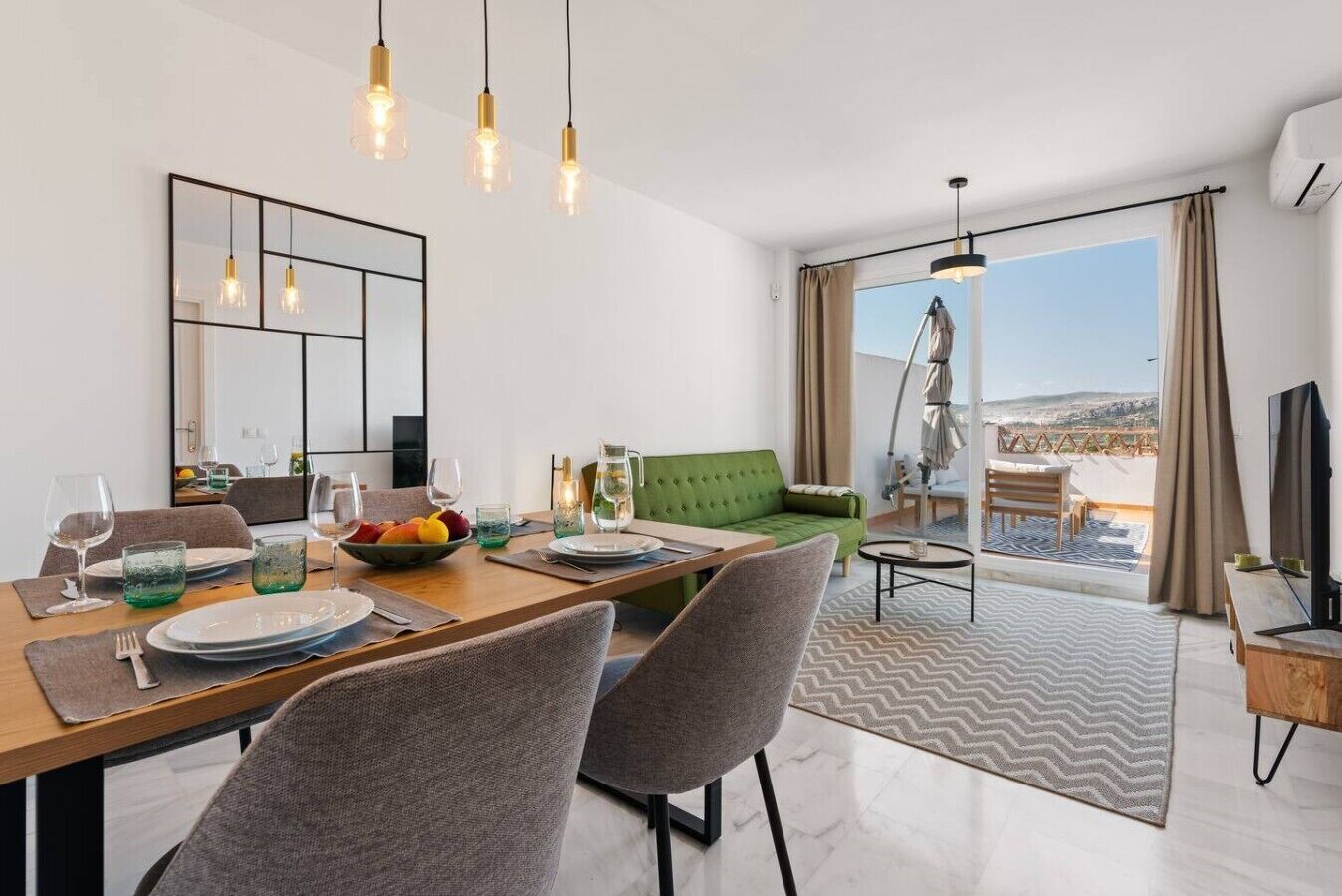 A modern dining area with a wooden table set for two, a green sofa, and a balcony with a view.