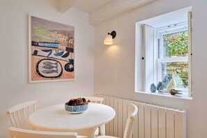 A dining area with a round table, white chairs, a bowl of food, a wall-mounted light, and a window with a view of greenery. - Refined Cornish Heritage Cottage for 3 (Penzance)