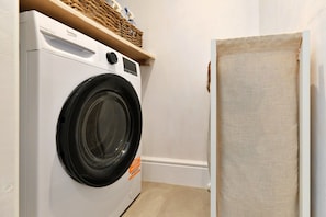 A compact laundry room with a front-loading washing machine, a wicker shelf above it, and a partially open door to another room. - Refined Cornish Heritage Cottage for 3 (Penzance)