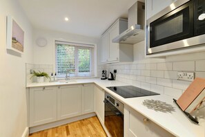A modern kitchen with white cabinetry, a built-in microwave, and a window with blinds. - Refined Cornish Heritage Cottage for 3 (Penzance)