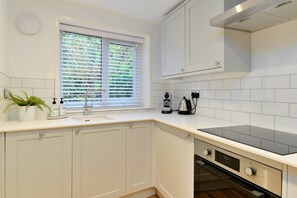 A modern kitchen with white cabinetry, a built-in oven, and a window with blinds. - Refined Cornish Heritage Cottage for 3 (Penzance)