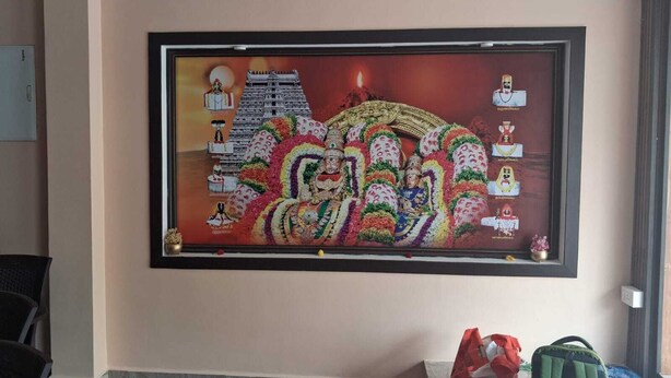 Obra enmarco que muestra una escena tradicional con figuras y una estructura de templo, colocada en una pared de una habitación con ventilador de techo y una silla.