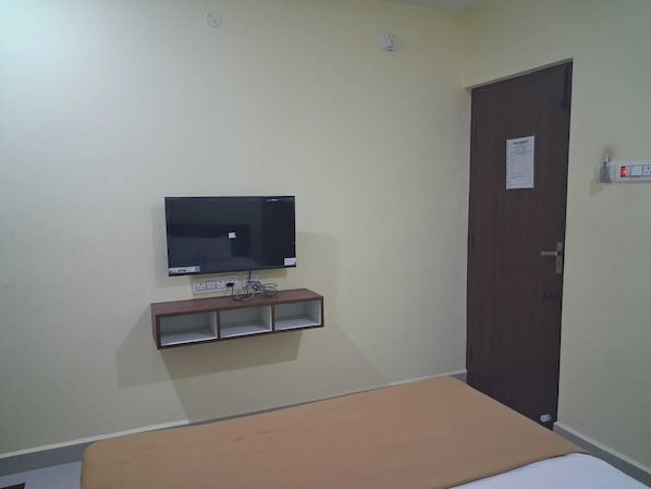A room with a bed, a television mounted on the wall, a door with a notice, and electrical outlets. - SAKTHI RESIDENCY (Tiruvannamalai)