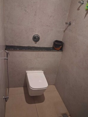 A small, enclosed shower stall with a white toilet, a metal showerhead, and a trash bin. - SAKTHI RESIDENCY (Tiruvannamalai)