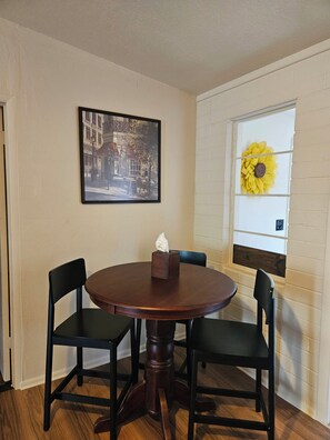 A dining area with a round table, two chairs, a framed picture on the wall, and a sunflower decoration on the window.