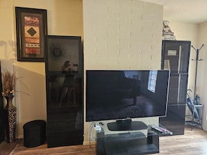 Una sala de estar con un televisor de pantalla plana grande sobre un pedestal de vidrio, un cuadro en la pared y una mesita con un jarrón de plantas secas.
