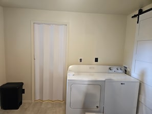 A laundry room with a washer and dryer, a trash bin, and a door with a striped curtain. - Red Rock Rendezvous - Steps Away From Uptown Sedona Shops & Dining (Sedona)