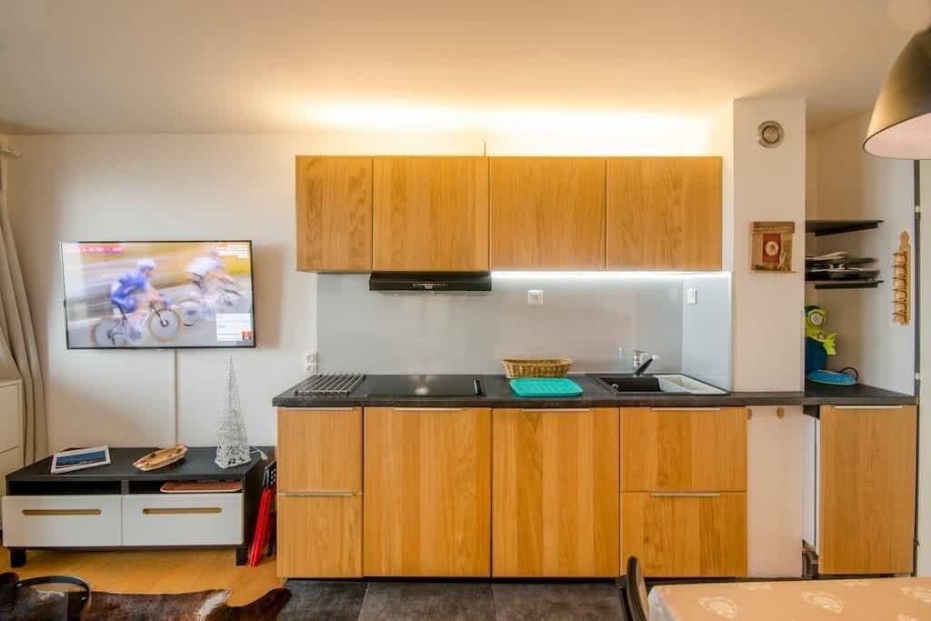 Eine kompakte Küche mit Holzausstattung, einer schwarzen Arbeitsplatte und einem an der Wand befestigten Fernseher.