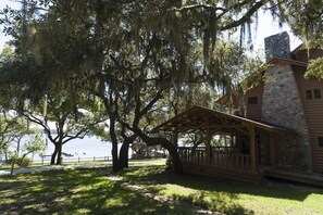 A wooden cabin with a stone chimney, surrounded by large trees and a body of water. - Sunnyside Villa - Beach Access, Fire Pit & Family Fun Near 30A, Sleeps 8 (Panama City Beach)