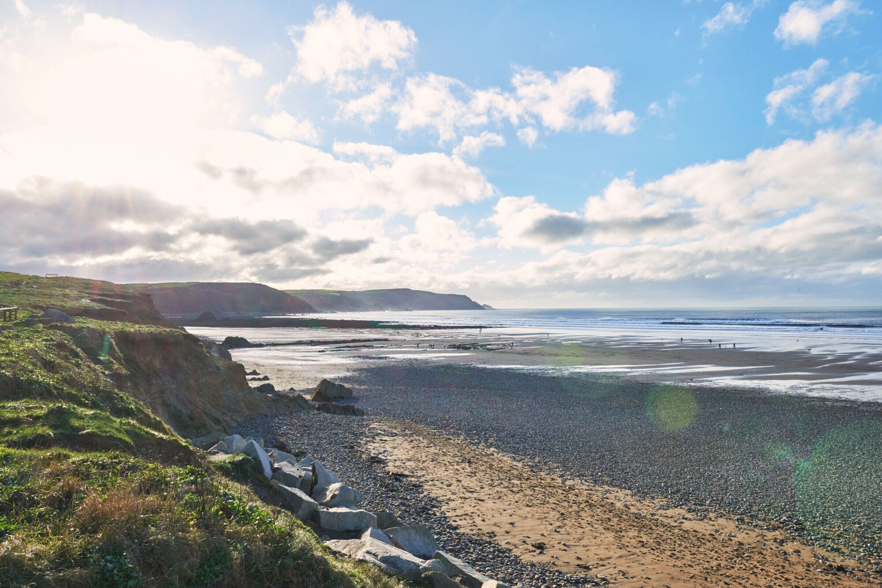 A coastal landscape with a pebbly beach, grassy cliffs, and a clear sky.