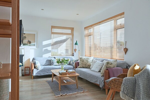 A living room with a sofa, a coffee table with books, and a window with blinds. - Coastal Family House for 8 in Cornwall (Bude)
