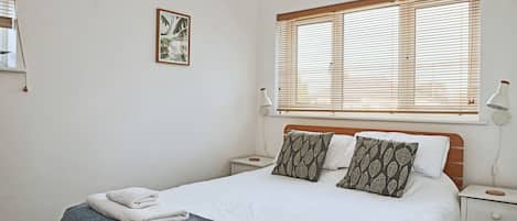 Un dormitorio con una cama, dos mesitas de noche, una ventana con persianas y una lámpara de pared, además de un cuadro enmarcado.