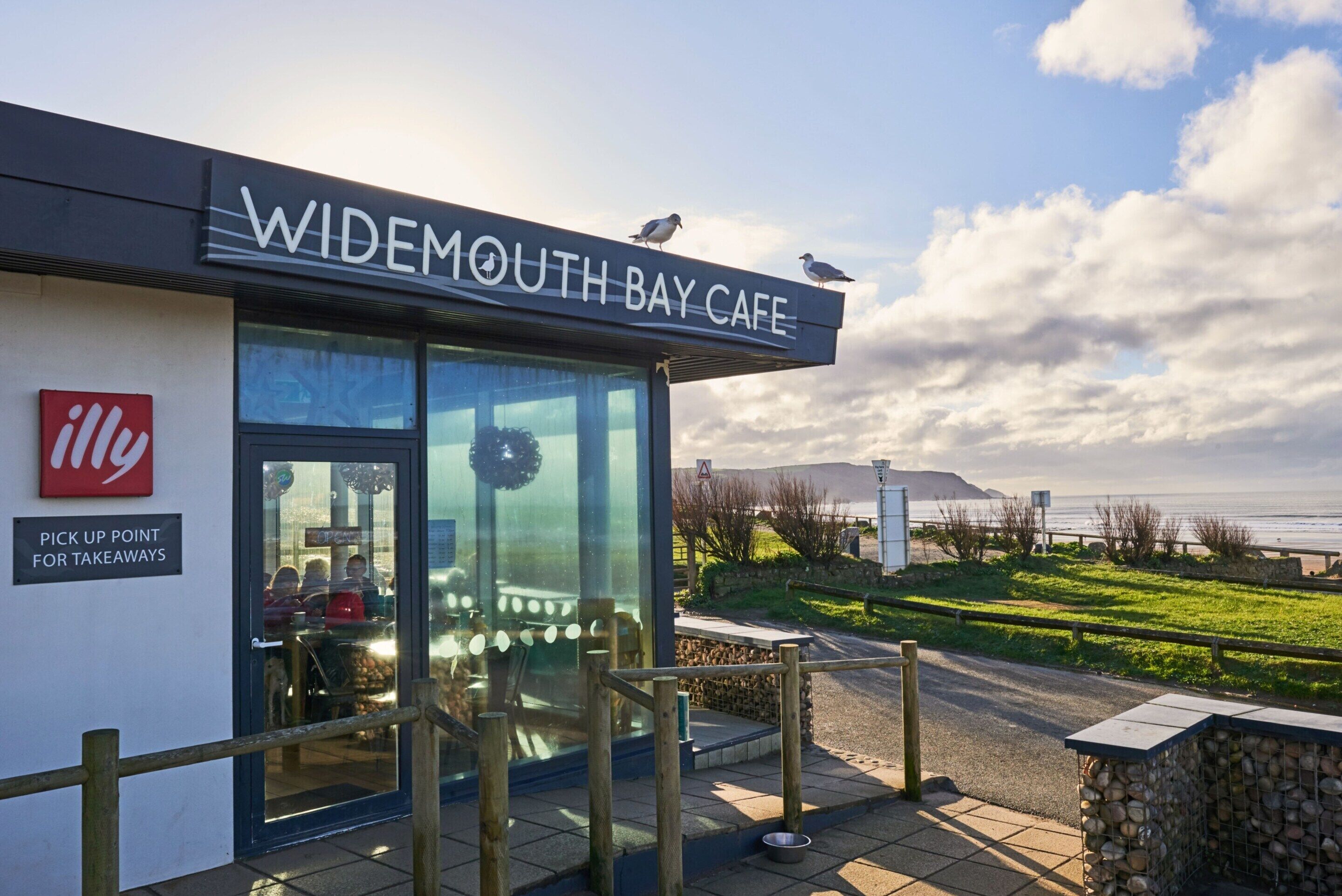 A waterfront cafe named 'Widemouth Bay Cafe' with a sign indicating a pick-up point for takeaways.