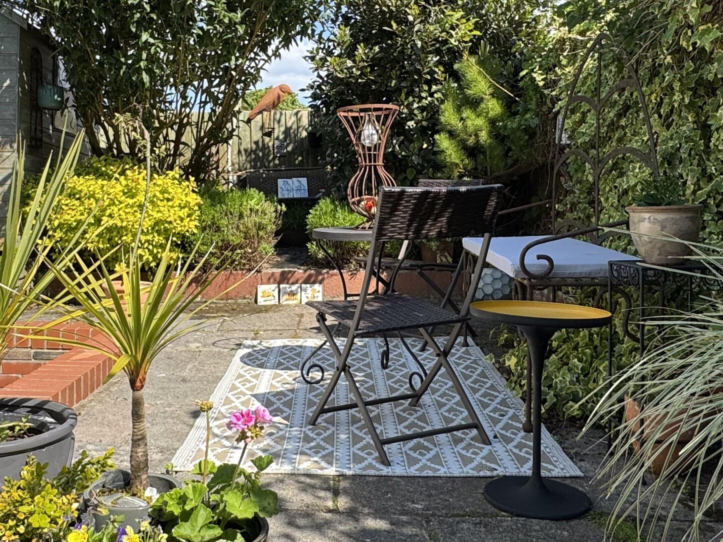 Une terrasse extérieure avec une table et des chaises en métal, entourée de plantes en pot et d’un tapis à motifs.