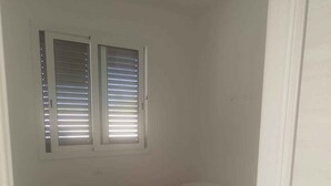 Une petite chambre avec un lit simple, une couvre-lit à motifs floraux verts, deux fenêtres équipées de stores et une porte blanche.
