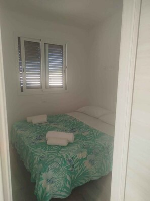 Ein kleines Zimmer mit einem Einzelbett, einer grün gemusterten Tagesdecke, zwei Fenstern mit Jalousien und einer weißen Tür.