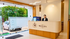 A reception desk with a logo, a receptionist, and a view of the outside. - Hotel Mewada (Ujjain)