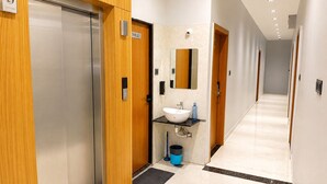 A modern hotel corridor with a wooden elevator, a sign indicating a toilet, a sink with a mirror, and a trash bin. - Hotel Mewada (Ujjain)
