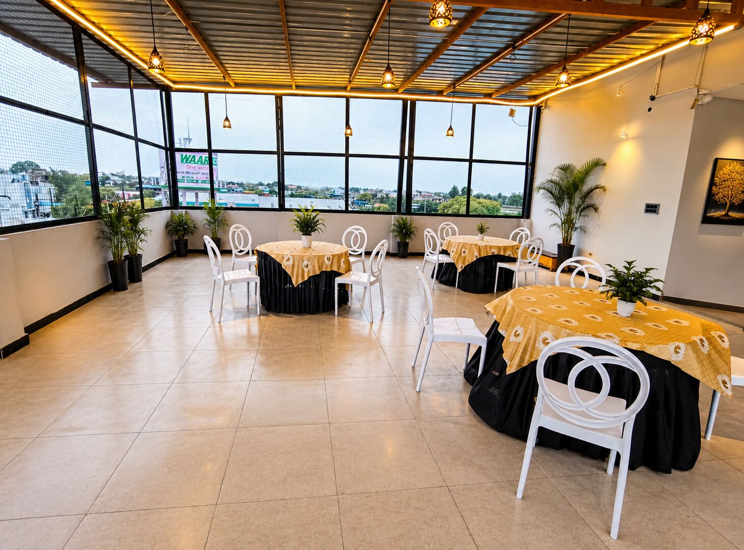 Um espaço interno amplo, com várias mesas redondas e cadeiras, grandes janelas que oferecem vista do exterior e luminárias pendentes.