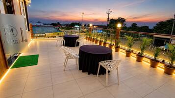 Una terraza en la azotea con mesas y sillas redondas, plantas en macetas y vistas de la ciudad al anochecer.