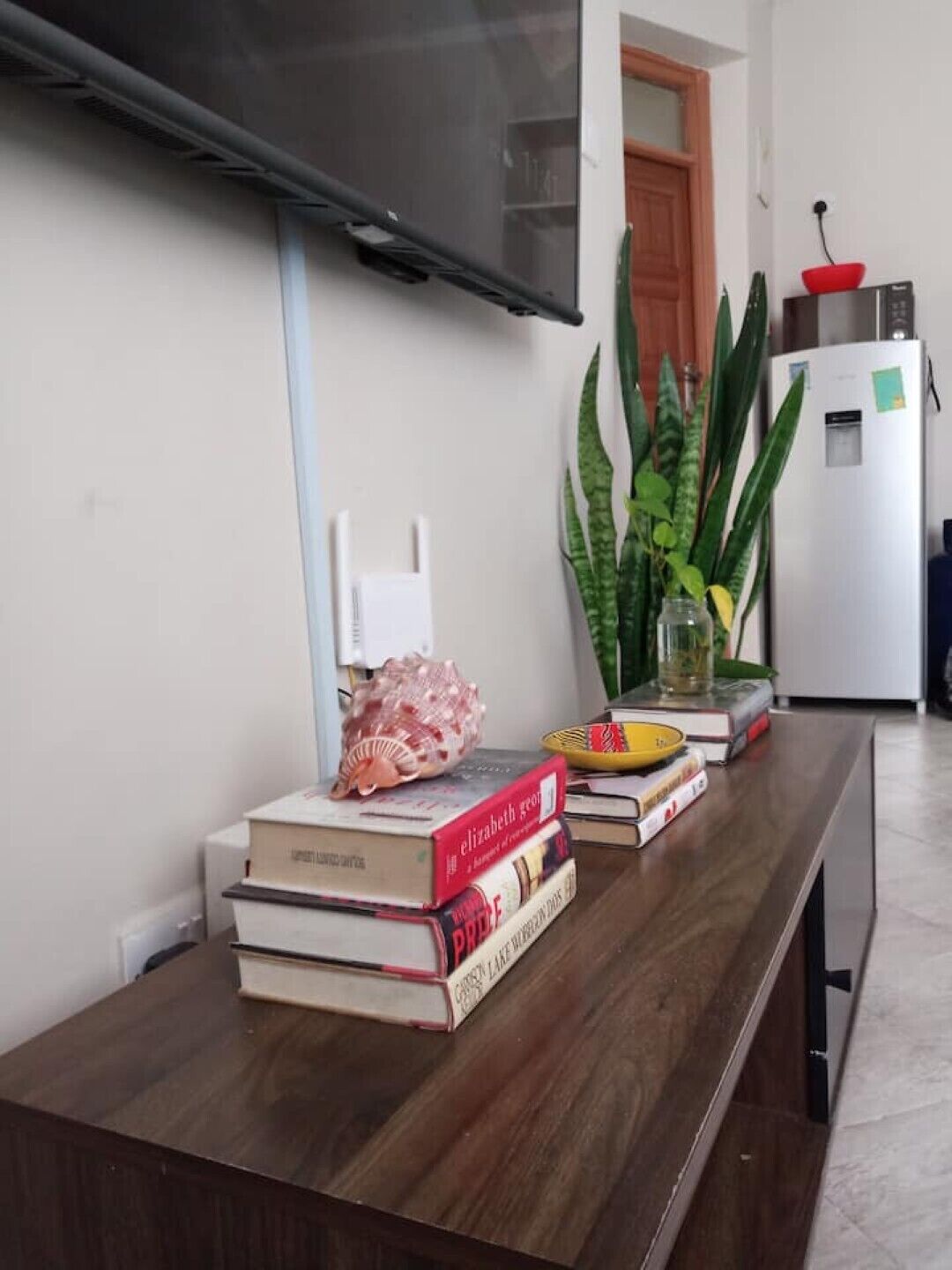 Une pièce avec une table en bois, des livres, un téléviseur, un réfrigérateur et une plante en pot.