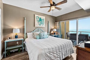 A bedroom with a bed, a ceiling fan, a nightstand, a lamp, a wall art piece, and a view of the ocean through a sliding door. - Emerald Isle Unit 1402 (Pensacola Beach)