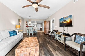 Un salon comprenant un canapé, une table basse et un téléviseur. Il y a également un coin repas avec des chaises et une table. La pièce est dotée d’un plancher en bois franc et d ’un ventilateur de plafond.