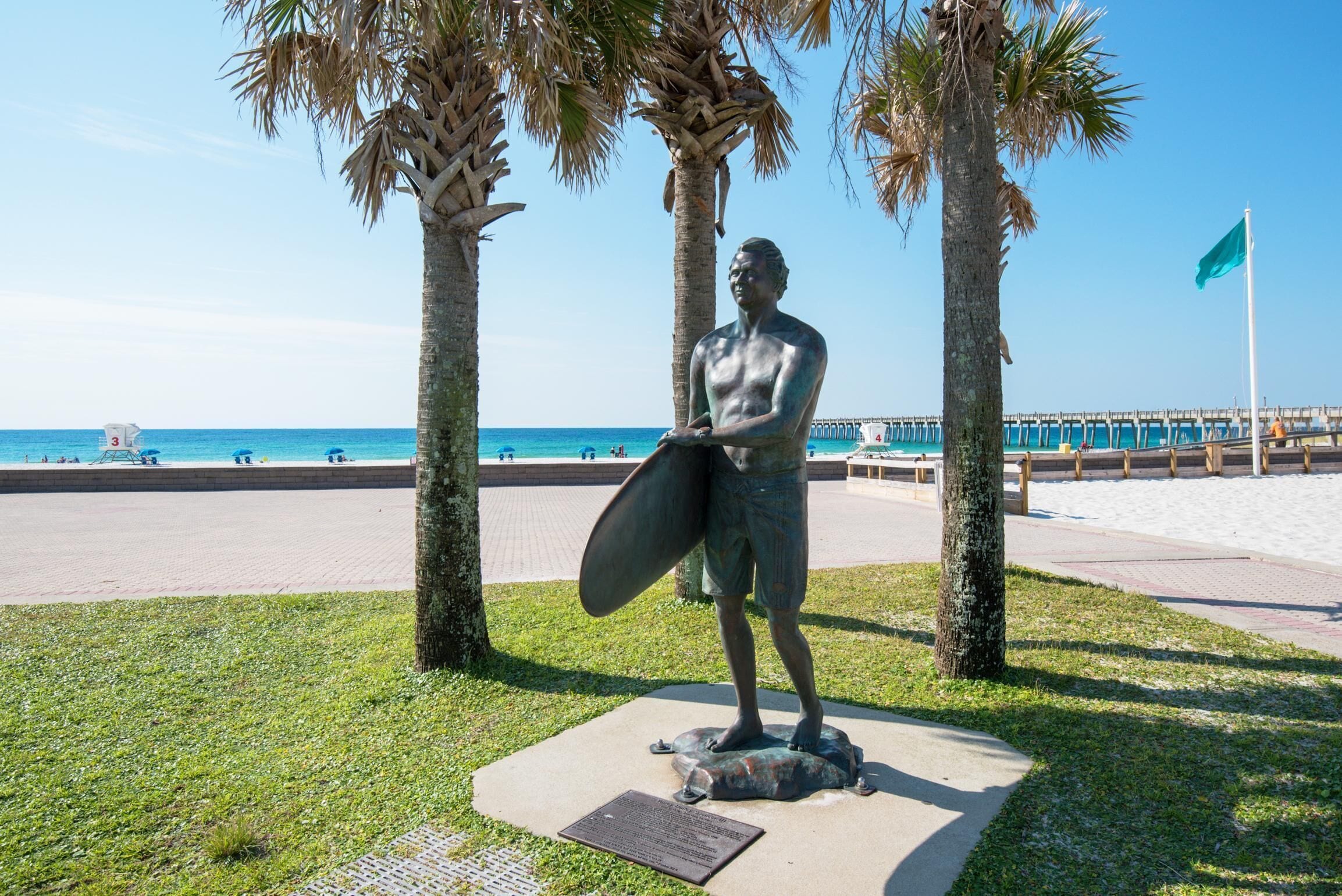 A statue of a man holding a surfboard on a pedestal, surrounded by palm trees, with a beach and pier in the background.