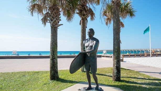 Estátua de um homem segurando uma prancha de surf em um pedestal, cercada por palmeiras, com uma praia e um píer ao fundo.