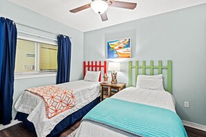 A bedroom with two beds, a ceiling fan, a window with curtains, and a painting on the wall. - Emerald Isle Unit 1402 (Pensacola Beach)
