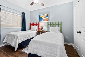 A bedroom with two beds, a ceiling fan, a painting, and a window with curtains. - Emerald Isle Unit 1402 (Pensacola Beach)