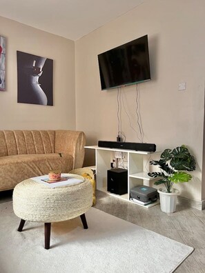 Ein Wohnzimmer mit einer Couch, einem an der Wand montierten Fernseher, einem kleinen Beistelltisch mit einer Pflanze und einem dekorativen Gegenstand auf dem Hocker.