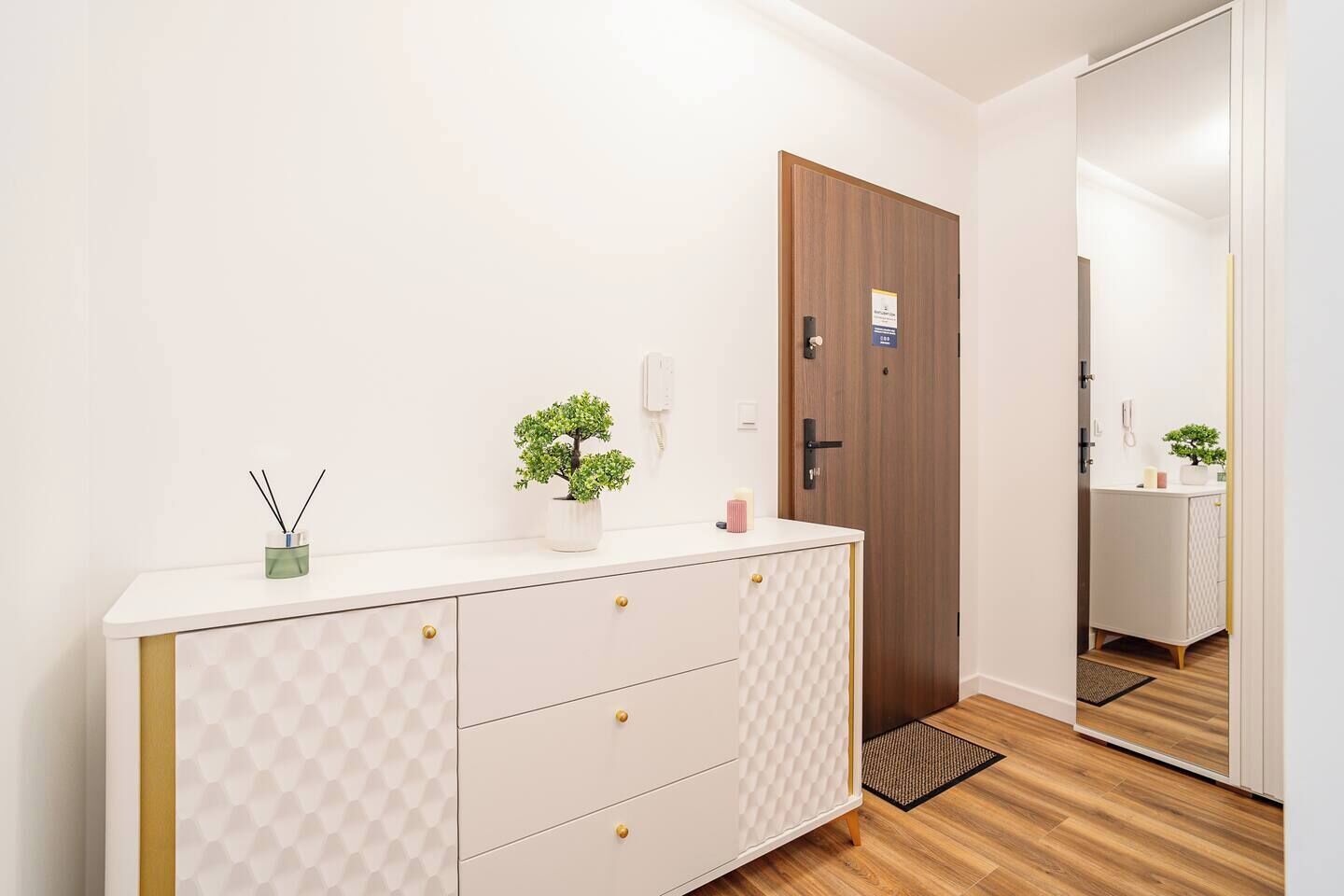 A hotel room with a white vanity, a wooden door, a small potted plant, and a mirror reflecting the room.
