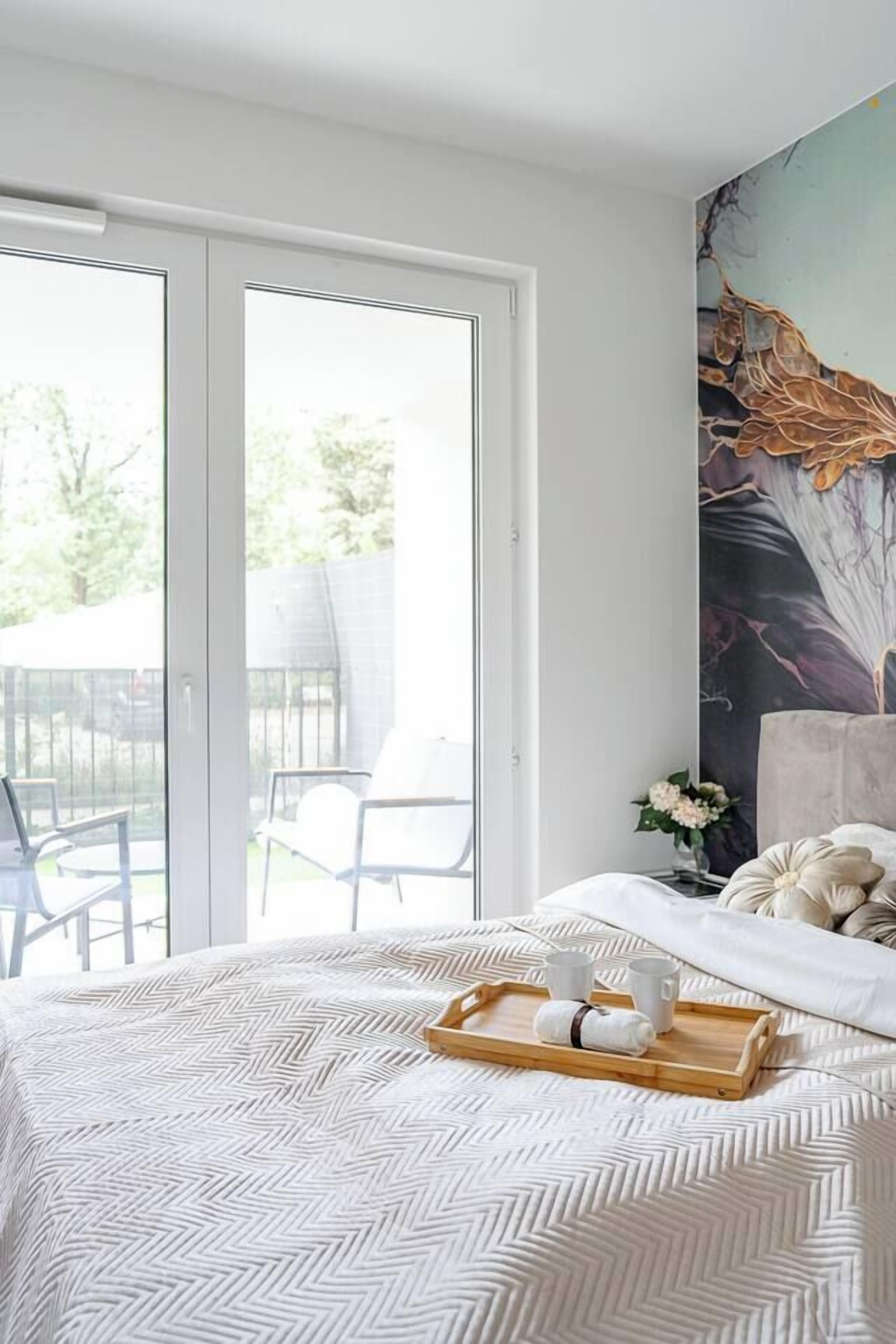 A bedroom with a bed, a tray with cups and a towel, a painting on the wall, and a view of the outdoors through a sliding door.