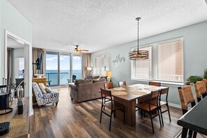 A spacious living area with a dining table, chairs, and a sofa. There is a balcony with a view of the ocean. - Summerwind West 804 (Navarre)