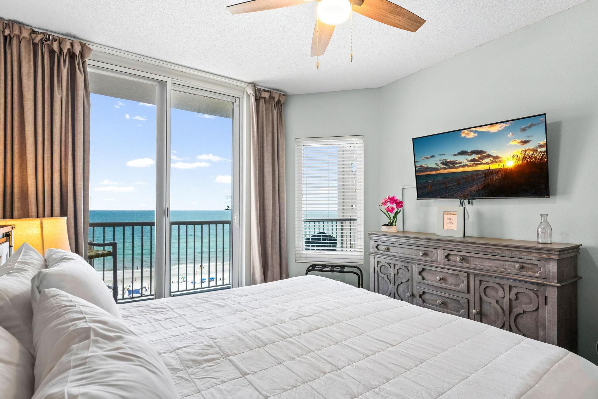 A hotel room with a large bed, a flat-screen TV, a balcony with a view of the sea, and a ceiling fan.