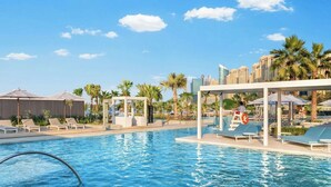 Ein Swimmingpool mit Liegestühlen und Sonnenschirmen, umgeben von Palmen und modernen Gebäuden.