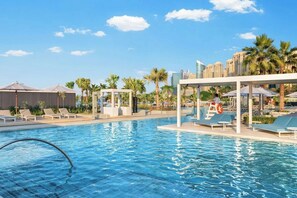 Ein Swimmingpool mit Liegestühlen und Sonnenschirmen, umgeben von Palmen und modernen Gebäuden.