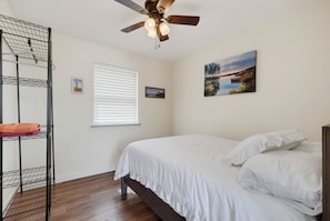 Une chambre à coucher avec un lit, un ventilateur de plafond, une fenêtre avec des stores et un tableau au mur.