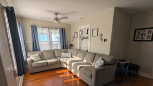 A living room with a sectional sofa, a ceiling fan, a rug, and a window with curtains. - Charming 2-bedroom 1935 bungalow in downtown New Port Richey (New Port Richey)
