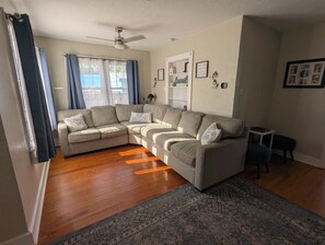 A living room with a sectional sofa, a ceiling fan, a rug, and a window with curtains. - Charming 2-bedroom 1935 bungalow in downtown New Port Richey (New Port Richey)
