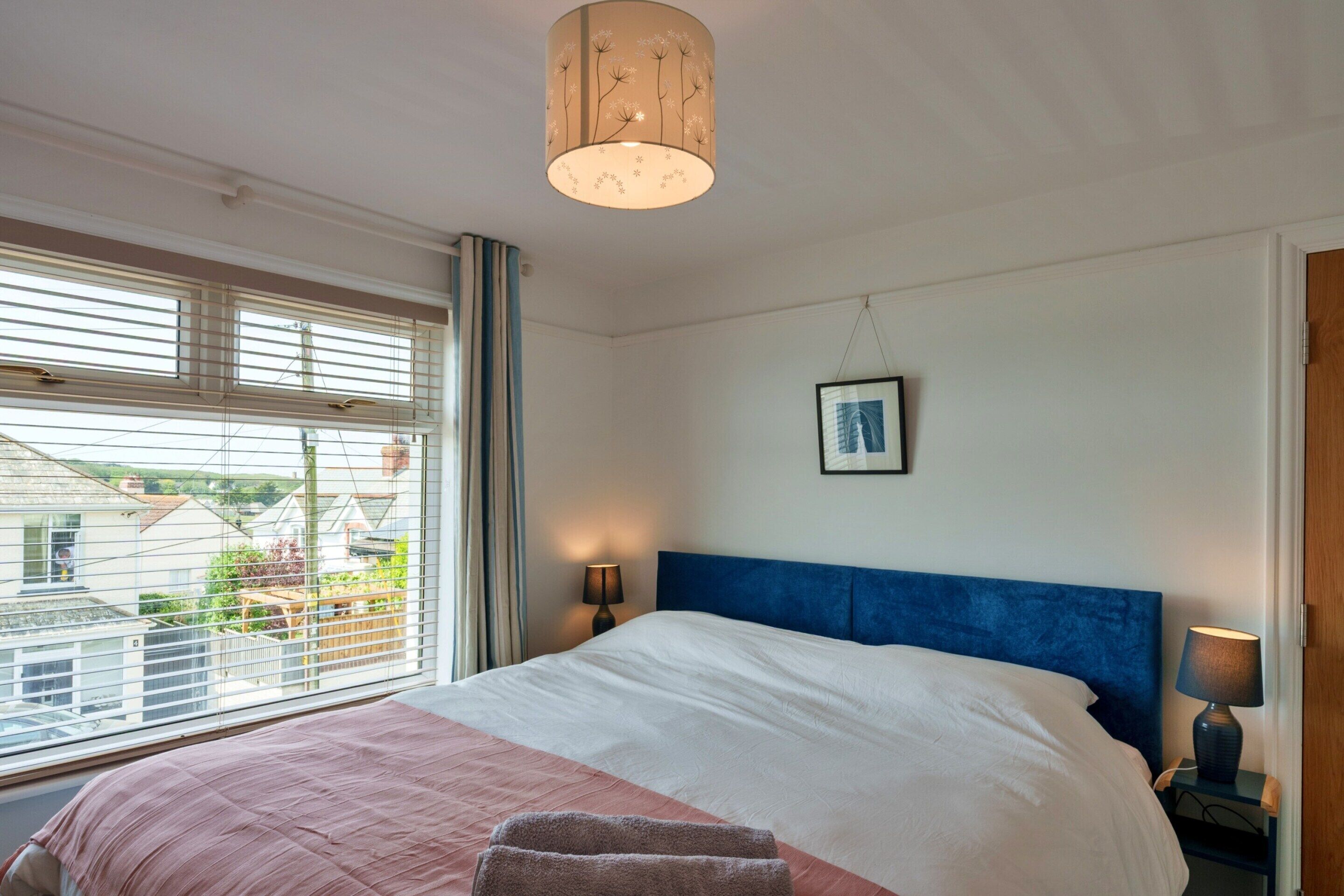 A bedroom with a bed, a bedside lamp, a window with blinds, and a view of houses outside.