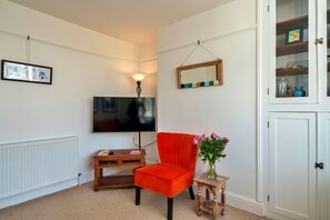 A living room with a television, a red armchair, a small table with a vase of flowers, and a cabinet with decorative items. - Peaceful Lochside Retreat for 4 (Bude)