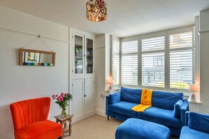 A room with a red chair, a blue sofa, a small table with a vase of flowers, and a window with blinds. - Peaceful Lochside Retreat for 4 (Bude)