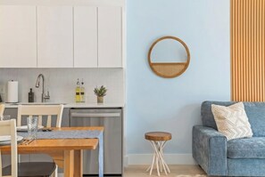 Eine moderne Küche mit weißen Schränken, einem Esstisch mit Stühlen, einem kleinen runden Holzschemel und einem blauen Sofa mit Kissen.