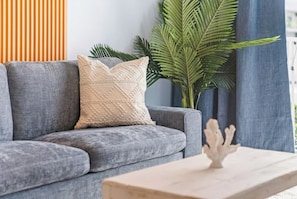 A grey sofa with a textured beige pillow, a small white decorative object on a table, and a large green plant in the background.