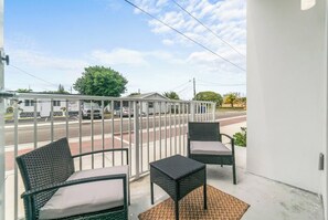 Ein Balkon mit zwei Korbstühlen und einem kleinen Tisch, mit Blick auf eine Straße, auf der Autos geparkt sind, und ein Gebäude im Hintergrund.