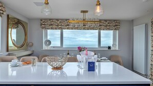 Ein Essbereich mit einem großen Tisch, mehreren Stühlen, einer Kronleuchter und Blick auf das Meer durch die Fenster.