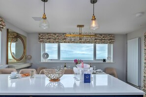 Ein Essbereich mit einem großen Tisch, mehreren Stühlen, einer Kronleuchter und Blick auf das Meer durch die Fenster.