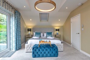 Un dormitorio con una cama grande, un reposapiés capitonado, una lámpara de techo circular y vistas a través de una ventana con cortinas.